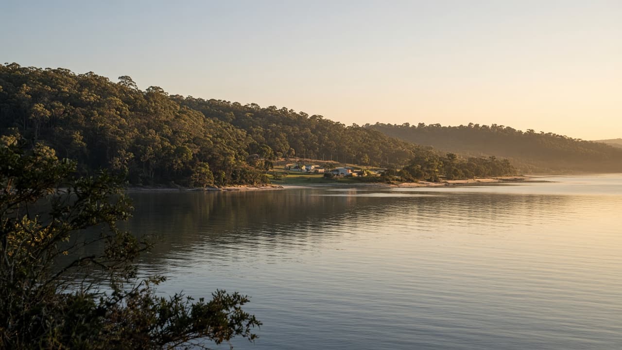 Generated illustration of a quiet forested bay settlement on calm deep water at golden hour, in the character of Boydtown on Twofold Bay.